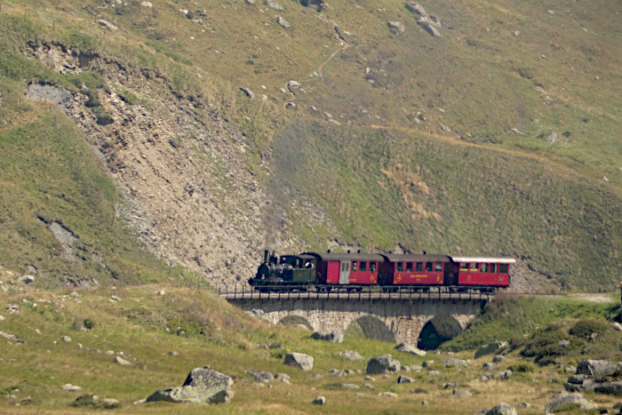 Die kleine HG2/3 exVZ Nr.6 WEISSHORN mit Zug Nr.139 Realp&rarr;Oberwald, von viel weiter oben an der Bahn aus fotografiert, wie der &bdquo;light&ldquo;-Zug mit den kurzen 3 Wagen gerade über den Steintafelsviadukt unten hinter Tiefenbach fährt, um 11:56h am 13.08.2026